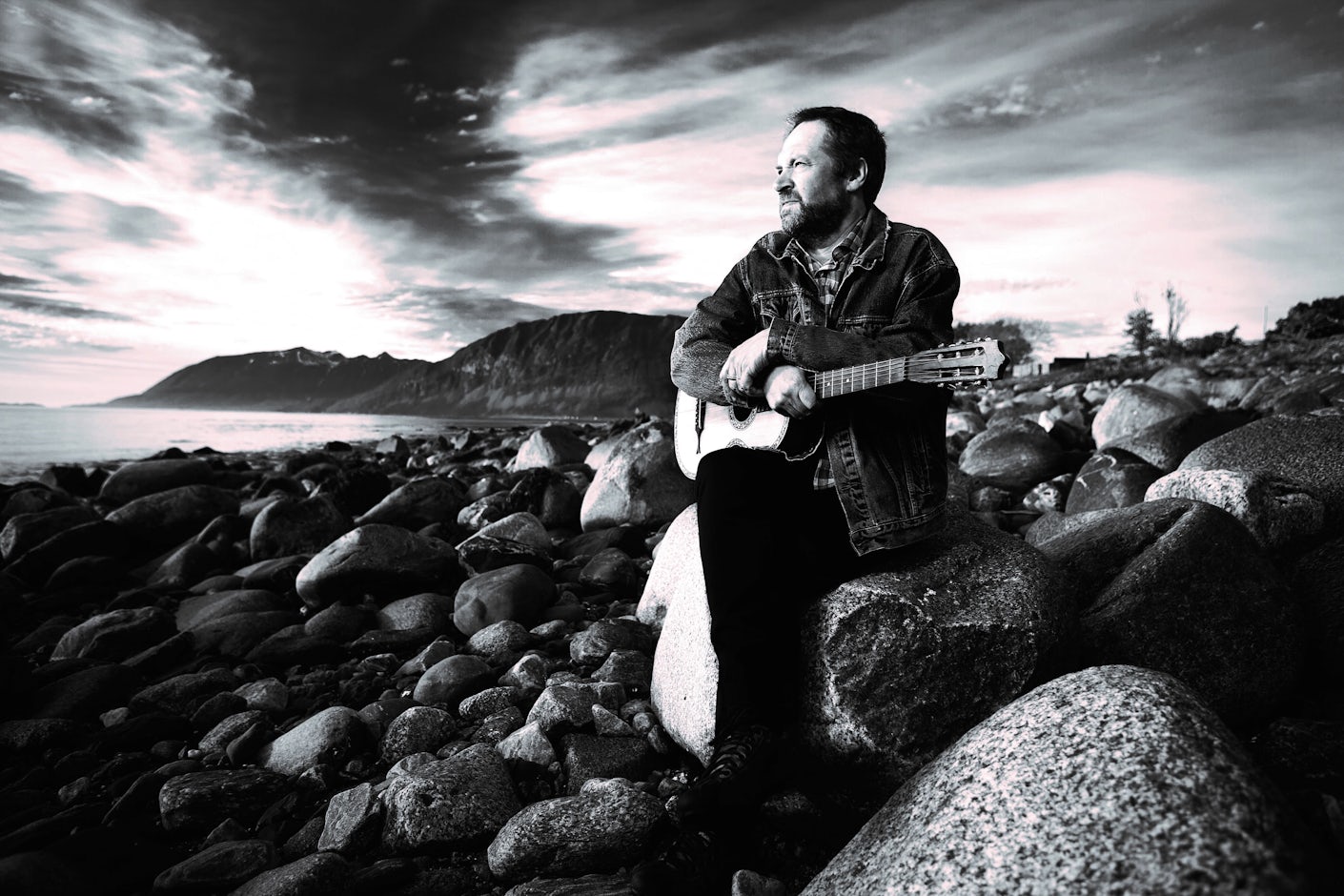 Sorthvitt bilde av en mann med gitar som ser utover havet-.