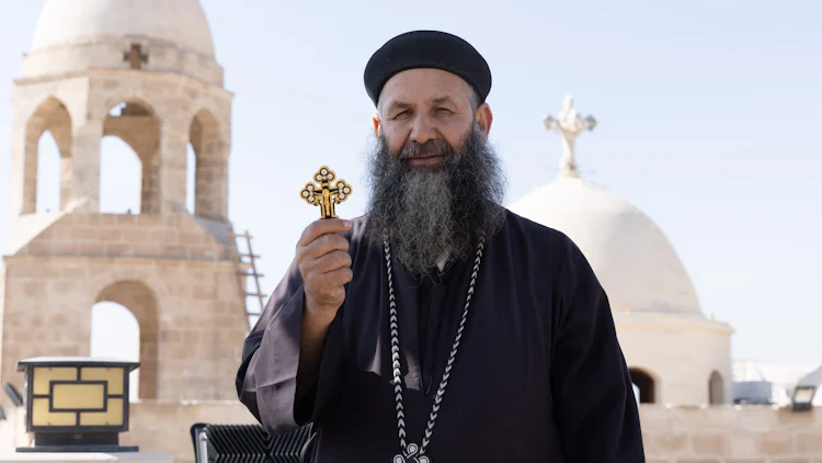 Ortodoks prest holder fram sitt kors foran deler av klosteranlegget i Minya