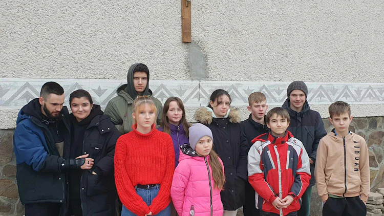 Barn og unge ved barnehjemmet samlet utendørs under et kors på husveggen.