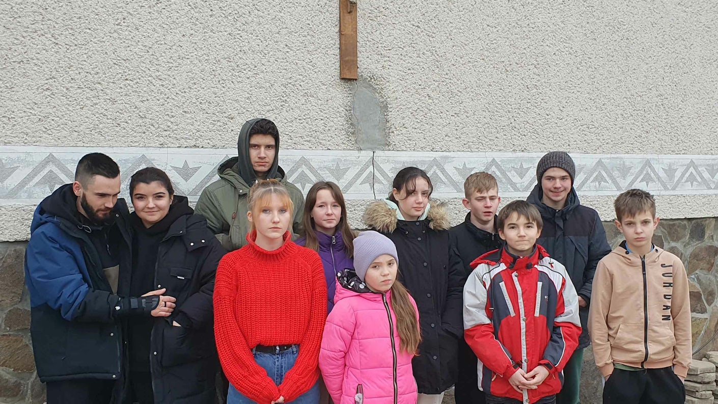 Barn og unge ved barnehjemmet samlet utendørs under et kors på husveggen.