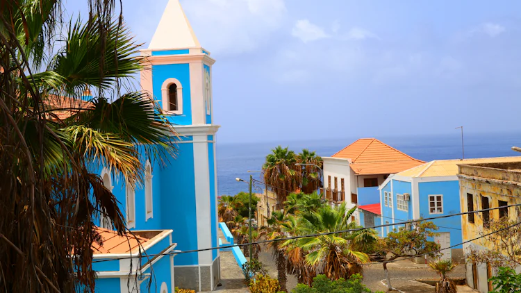 Gatebilde fra Kapp Verde med blåfarget kirke, byhus og palmetrær langs en gate som strekker seg mot havet i horisonten.